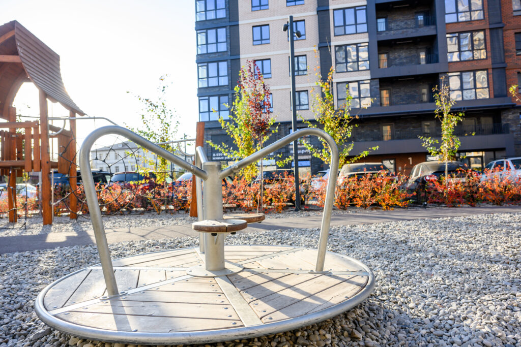A More Proactive Approach to Managing Risk in Communal Play Areas A metal merry-go-round sits on gravel in a playground, surrounded by colorful plants and a modern apartment building in the background
